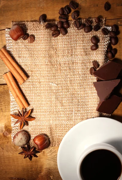 Coffee cup and beans, cinnamon sticks, nuts and chocolate on sacking on woo — Stock Photo, Image