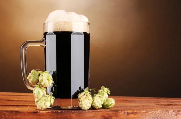 Cerveza oscura en una taza y lúpulo verde sobre una mesa de madera sobre fondo marrón — Foto de Stock