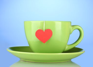 Green cup with saucer and tea bag with red heart-shaped label on blue backg clipart