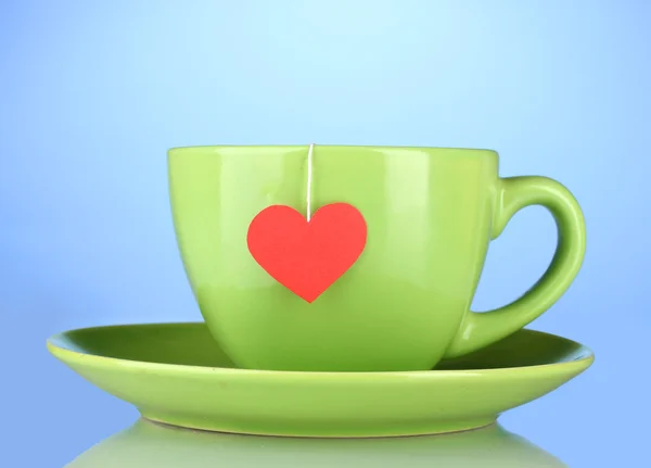 Green cup with saucer and tea bag with red heart-shaped label on blue backg — Stock Photo, Image
