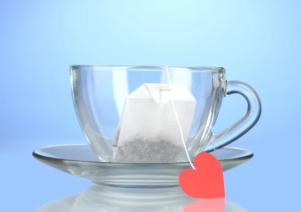 Glassy cup with saucer and tea bag with red heart-shaped label on blue back — Stock Photo, Image