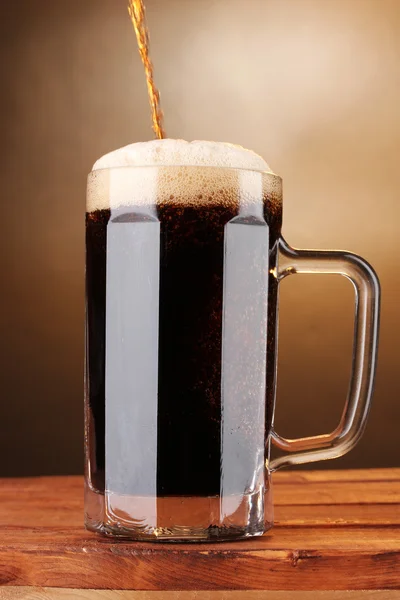 Tmavé pivo v hrnku na dřevěný stůl na hnědé pozadí — Stock fotografie