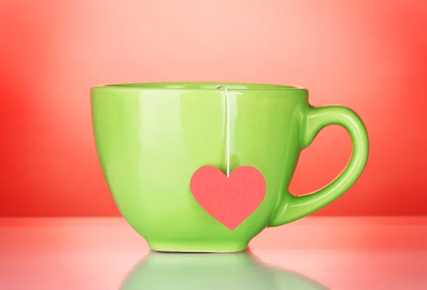 Green cup and tea bag with red heart-shaped label on red background — Stock Photo, Image