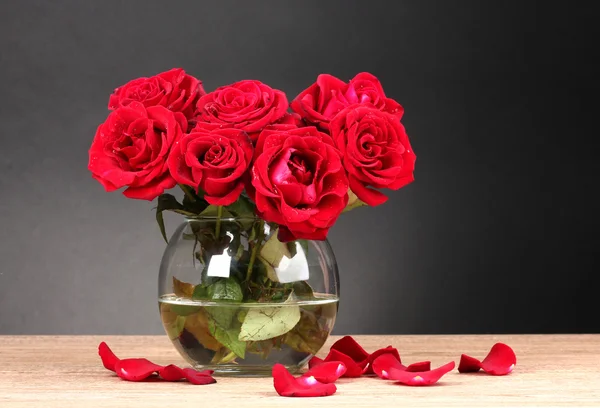 stock image Beautiful red roses in vase on wooden table on gray background