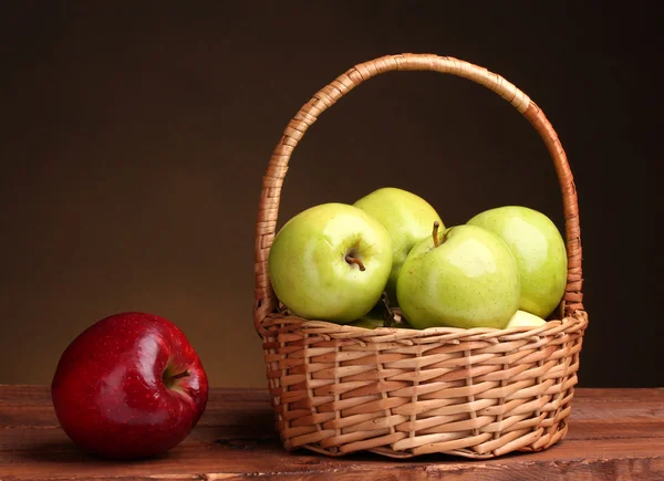 Soczyste zielone jabłka w kosz i czerwone jabłko na drewnianym stole na brązowy backgro — Zdjęcie stockowe