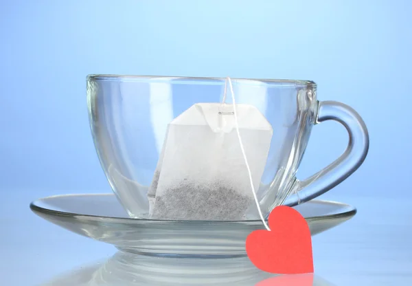 Glassy cup with saucer and tea bag with red heart-shaped label on blue back — Stock Photo, Image