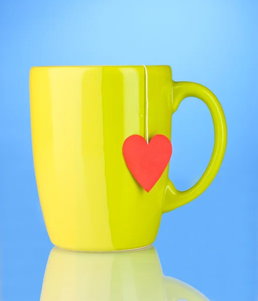 Green cup and tea bag with red heart-shaped label on blue background — Stock Photo, Image