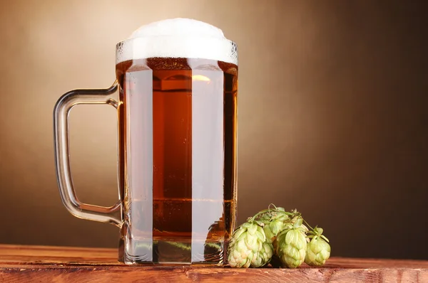 Jarra de cerveza y lúpulo verde sobre mesa de madera sobre fondo marrón —  Fotos de Stock