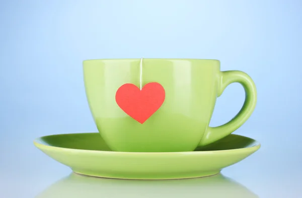 Green cup with saucer and tea bag with red heart-shaped label on blue backg — Stock Photo, Image