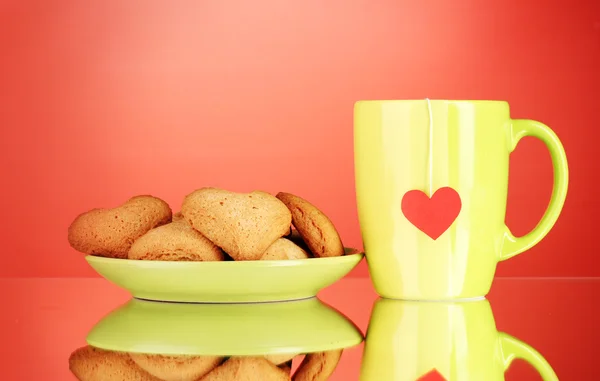 Cookies em forma de coração na placa e xícara com saco de chá no fundo vermelho — Fotografia de Stock
