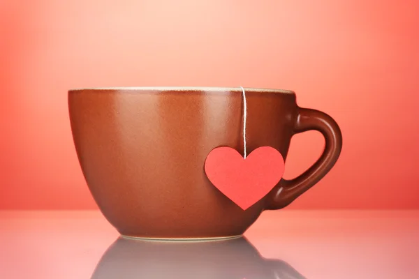 Brown cup and tea bag with red heart-shaped label on red background — Stock Photo, Image