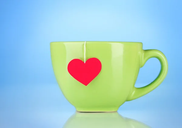 Green cup and tea bag with red heart-shaped label on blue background — Stock Photo, Image