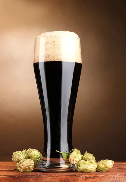 Birra scura in vetro e luppolo verde su tavolo in legno su sfondo marrone — Foto Stock
