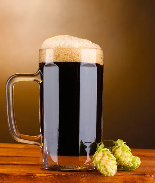 Cerveza oscura en una taza y lúpulo verde sobre una mesa de madera sobre fondo marrón —  Fotos de Stock