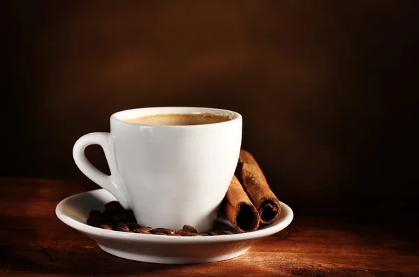 Чашка с кофе, корицей и кофейными зёрнами на деревянном столе на коричневом бэкгре — стоковое фото