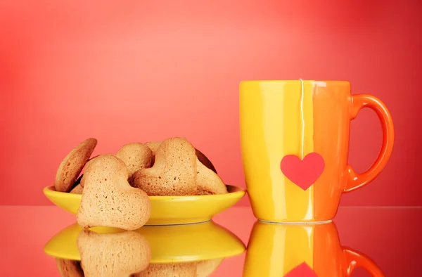 Heart-shaped cookies on yellow plate and cup with tea bag on red background — Stock Photo, Image