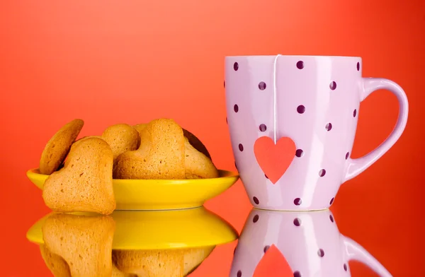 Çay poşeti kırmızı zemin üzerine sarı tabak ve bardak üzerinde kalp şeklinde kurabiye — Stok fotoğraf