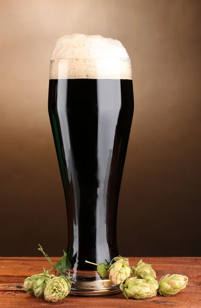 Dunkles Bier im Glas und grüner Hopfen auf Holztisch auf braunem Hintergrund — Stockfoto