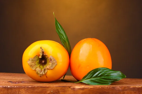 Two appetizing persimmons on wooden table on brown background — Stock Photo, Image