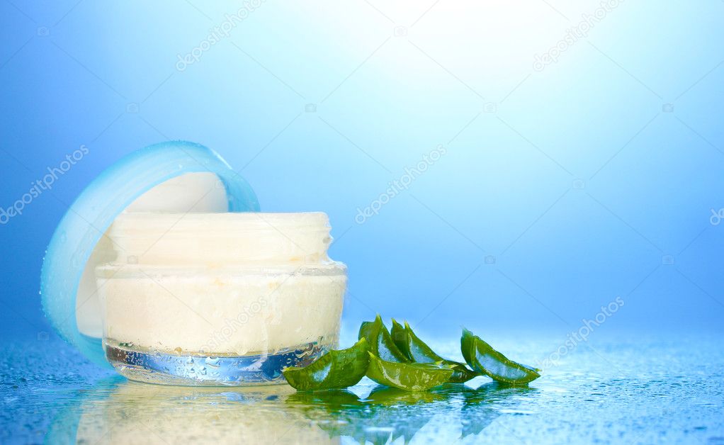 Opened glass jar of cream and aloe on blue background with water droplets