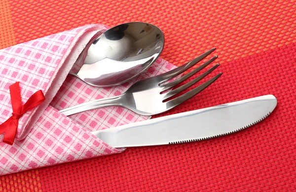 Tenedor, cuchara y cuchillo en un paño a cuadros con un lazo sobre un mantel rojo —  Fotos de Stock