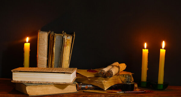 Old books, scrolls, ink pen inkwell and candles on wooden table on brown ba