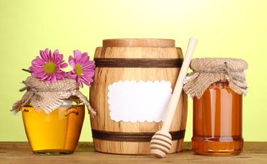 Sweet honey in jars and barrel with drizzler on wooden table on green background