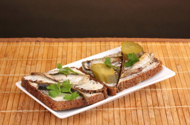 Tasty sandwiches with sprats on plate on wooden mat on brown background clipart