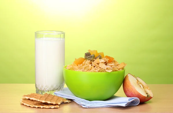 Tasty cornflakes in green bowl, apple and glass of milk on wooden table on green background — Stock Photo, Image