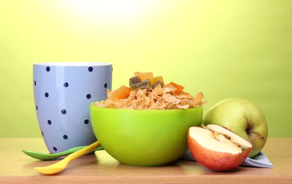 Tasty cornflakes in green bowl, apples and glass of milk on wooden table on green background — Stock Photo, Image