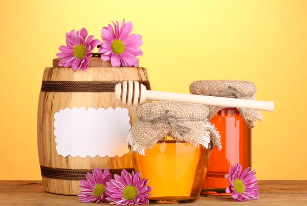 Süßer Honig in Gläsern und Fässern mit Nieselregen auf Holztisch auf gelbem Hintergrund — Stockfoto