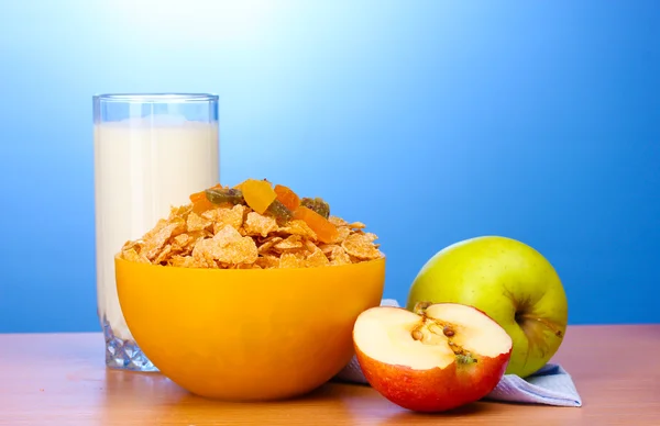 Välsmakande cornflakes i gul skål, äpplen och glas mjölk på träbord på blå bakgrund — Stockfoto