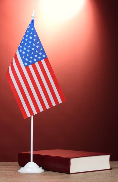 Americká vlajka na stojanu a kniha o dřevěný stůl na červeném pozadí — Stock fotografie