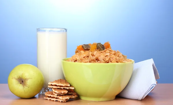Flocos de milho saborosos em tigela verde, maçãs e vidro de leite na mesa de madeira no fundo azul — Fotografia de Stock