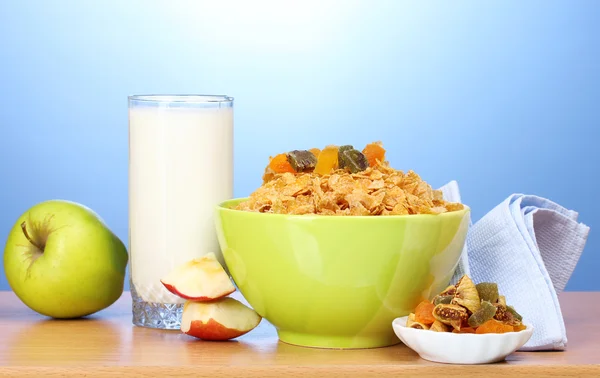 Chutné kukuřičné lupínky v zelené prohlubni, jablka a sklenici mléka na dřevěný stůl na modrém pozadí — Stock fotografie