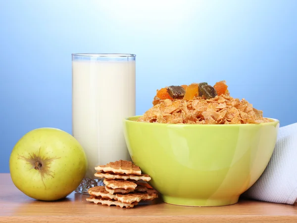 Välsmakande cornflakes i grön skål, äpplen och glas mjölk på träbord på blå bakgrund — Stockfoto