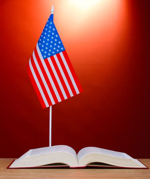 Bandeira americana no carrinho e livro sobre mesa de madeira no fundo vermelho — Fotografia de Stock