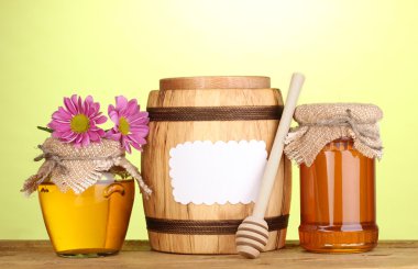 Sweet honey in jars and barrel with drizzler on wooden table on green background