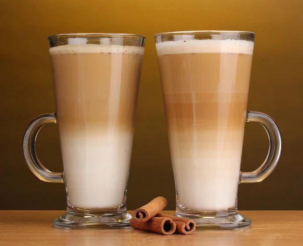 Pachnące? Unduli latte kubki szklane i cynamon na drewnianym stole na brązowym tle — Zdjęcie stockowe
