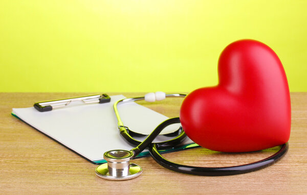 Medical stethoscope with clipboard and heart on wooden table on green background