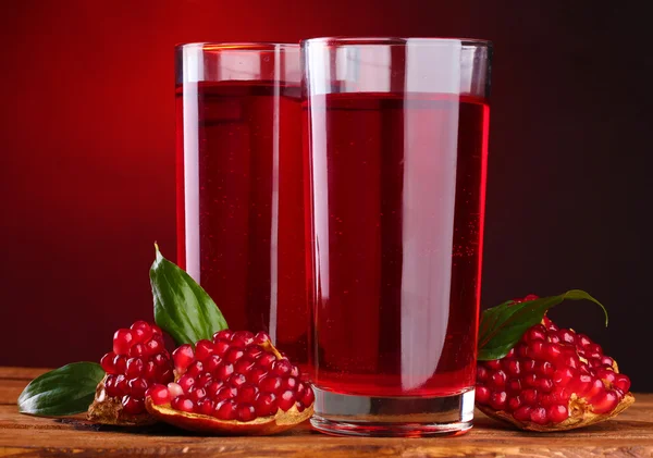 Dojrzałe pomergranate i szklanki soku na drewnianym stole na czerwonym tle — Zdjęcie stockowe