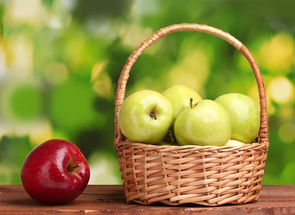 Manzanas verdes jugosas en cesta y manzana roja sobre mesa de madera sobre fondo verde —  Fotos de Stock