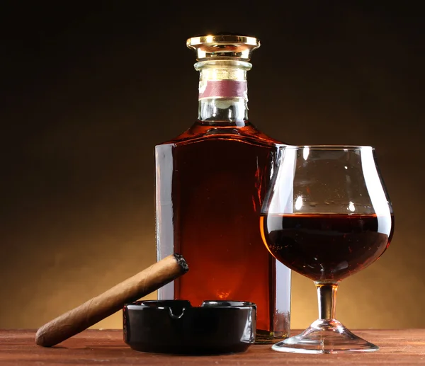 Botol dan gelas brendi dan cerutu di atas meja kayu dengan latar belakang coklat — Stok Foto