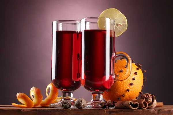 Vin brulè nei bicchieri, spezie e arancia su tavolo di legno su sfondo viola — Foto Stock