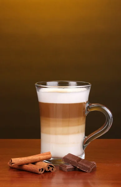 Latte au cappuccino parfumé dans une tasse en verre avec cannelle et chocolat sur table en bois sur fond marron — Photo