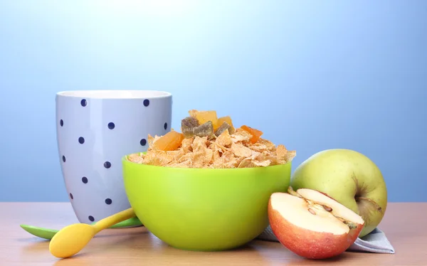Chutné kukuřičné lupínky v zelené prohlubni, jablka a sklenici mléka na dřevěný stůl na modrém pozadí — Stock fotografie