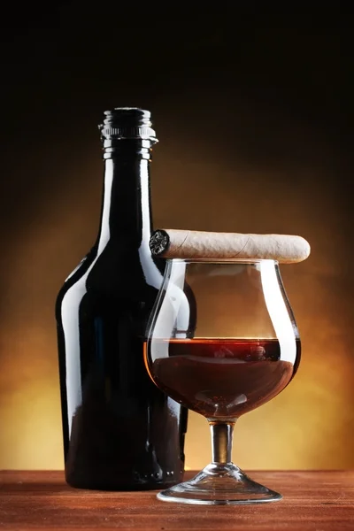 Botella y vaso de brandy y cigarro sobre mesa de madera sobre fondo marrón — Foto de Stock