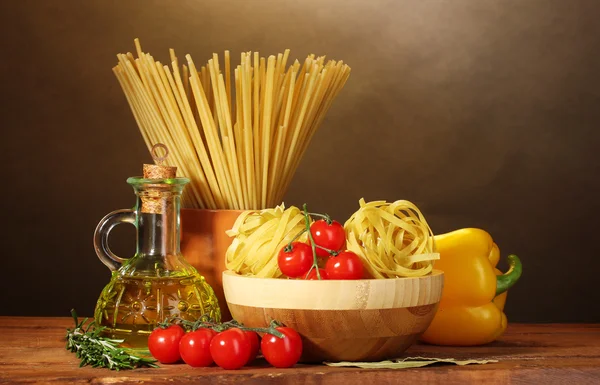 Špagety, nudle v misce, sklenici ropy a zeleniny na dřevěný stůl na hnědé pozadí — Stock fotografie
