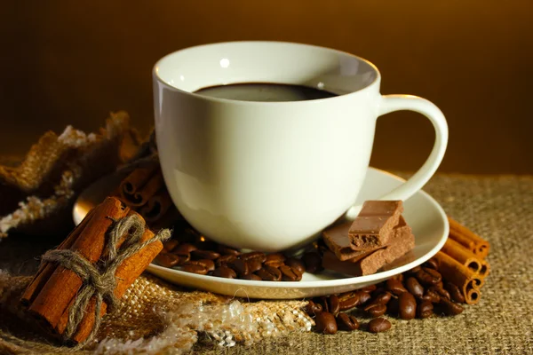 Kopp kaffe och bönor, kanelstänger och choklad på säckväv på brun bakgrund — Stockfoto