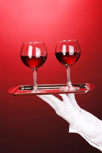 Main dans le gant tenant plateau d'argent avec verres à vin sur fond rouge — Photo
